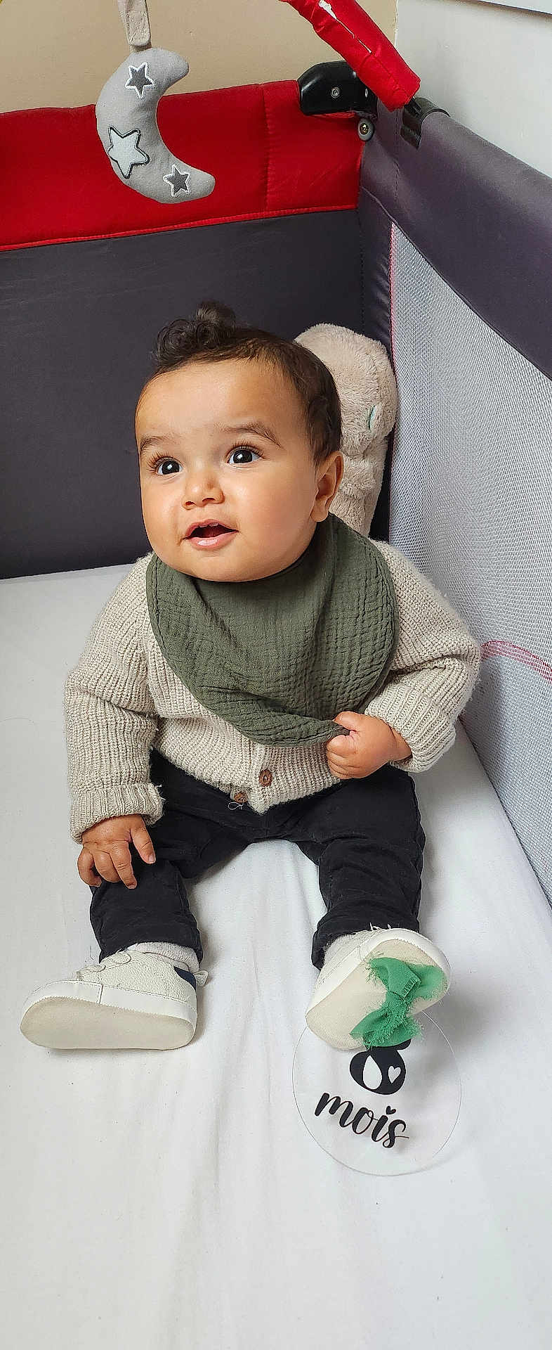 Maëlo participe au concours pour gagner de l'argent avec cette photo : baby, bib, big_eyes, brown_hair, child, cozy, crib, cute, hand, infant, milestone_disc, mobile_toy, moon_toy, playpen, plush_toy, portrait, sitting, smiling, sweater, white_shoes