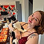 woman, cat, indoor, smiling, bow_tie, tattoo, red_dress, watch, bracelet, chair, table, geometric_wall, decor, floor, plant, television, holiday_decor, happy, person, holding