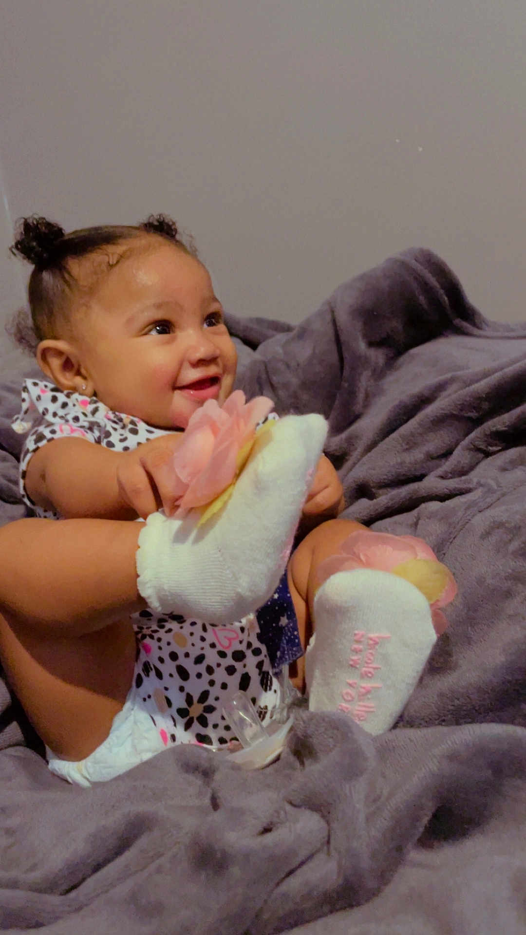Keylani joined the competition — help win amazing prizes! baby, infant, smiling, blanket, socks, pink_flower, pacifier, onesie, diaper, hair_buns, earrings, cozy, portrait, indoor, happy, sitting, soft_texture, gray_blanket, foot_focus, cute