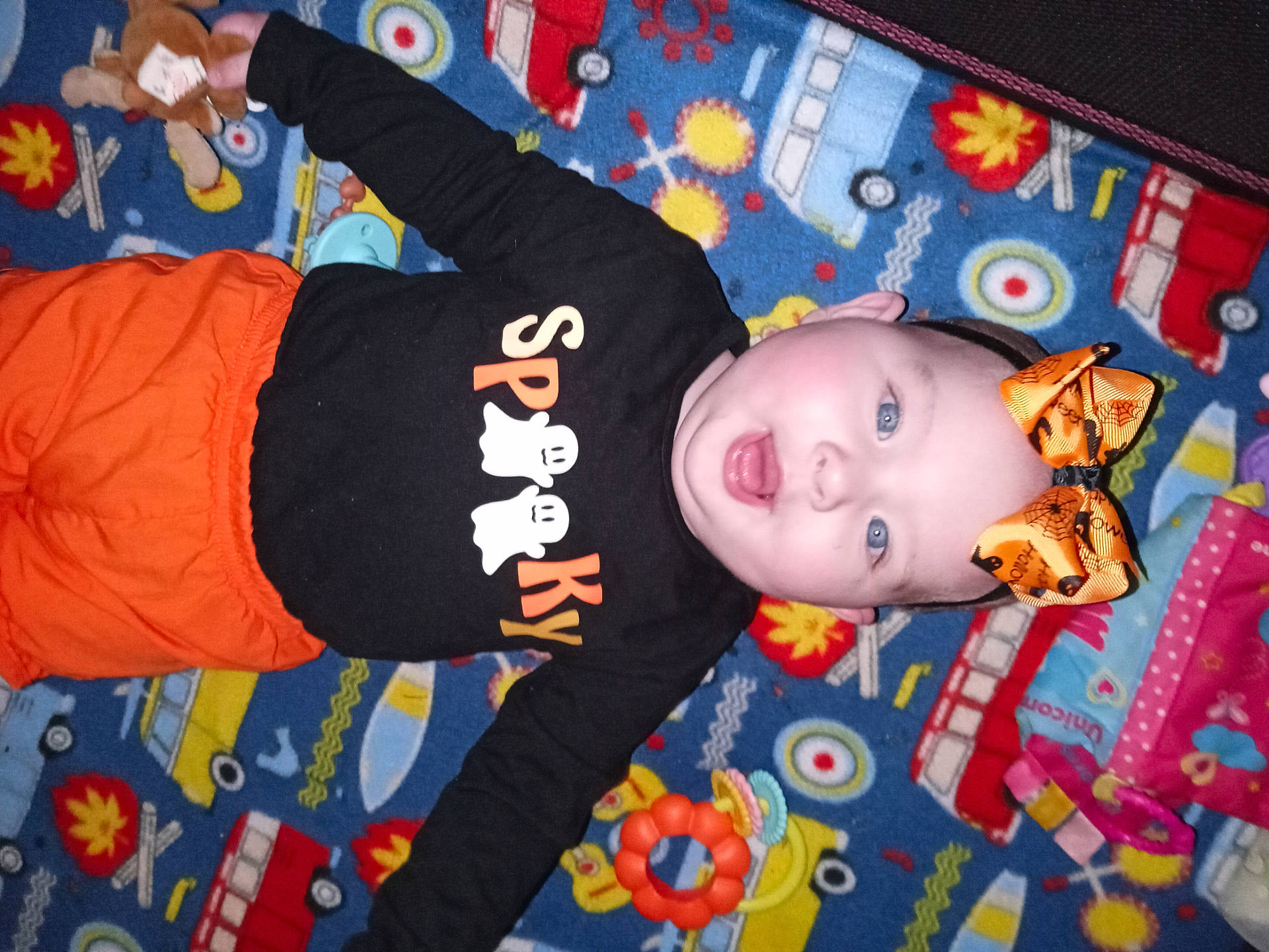 Lilianne is registered to the contest to win money with this photo: baby, black_shirt, blue_eyes, bow, car_pattern, child, colorful, cute, face, flower_pattern, ghosts, halloween, happy, headband, indoor, infant, orange_pants, play_mat, smiling, toy