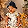 Noah Sotolongo is registered to the contest to win money with this photo: baby, child, plush_toy, stuffed_animal, lion_king, simba, hat, bucket_hat, romper, suspenders, striped_clothing, indoor, carpet, fluffy_rug, plant, wooden_crate, decor, cute, portrait, sitting