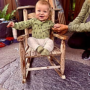 Owen is registered to the contest to win money with this photo: baby, child, rocking_chair, green_shirt, socks, hand_holding, smiling, indoor, wooden_chair, person, adult_hand, floor, plant, flag, carpet, furniture, happy, portrait, cozy, support