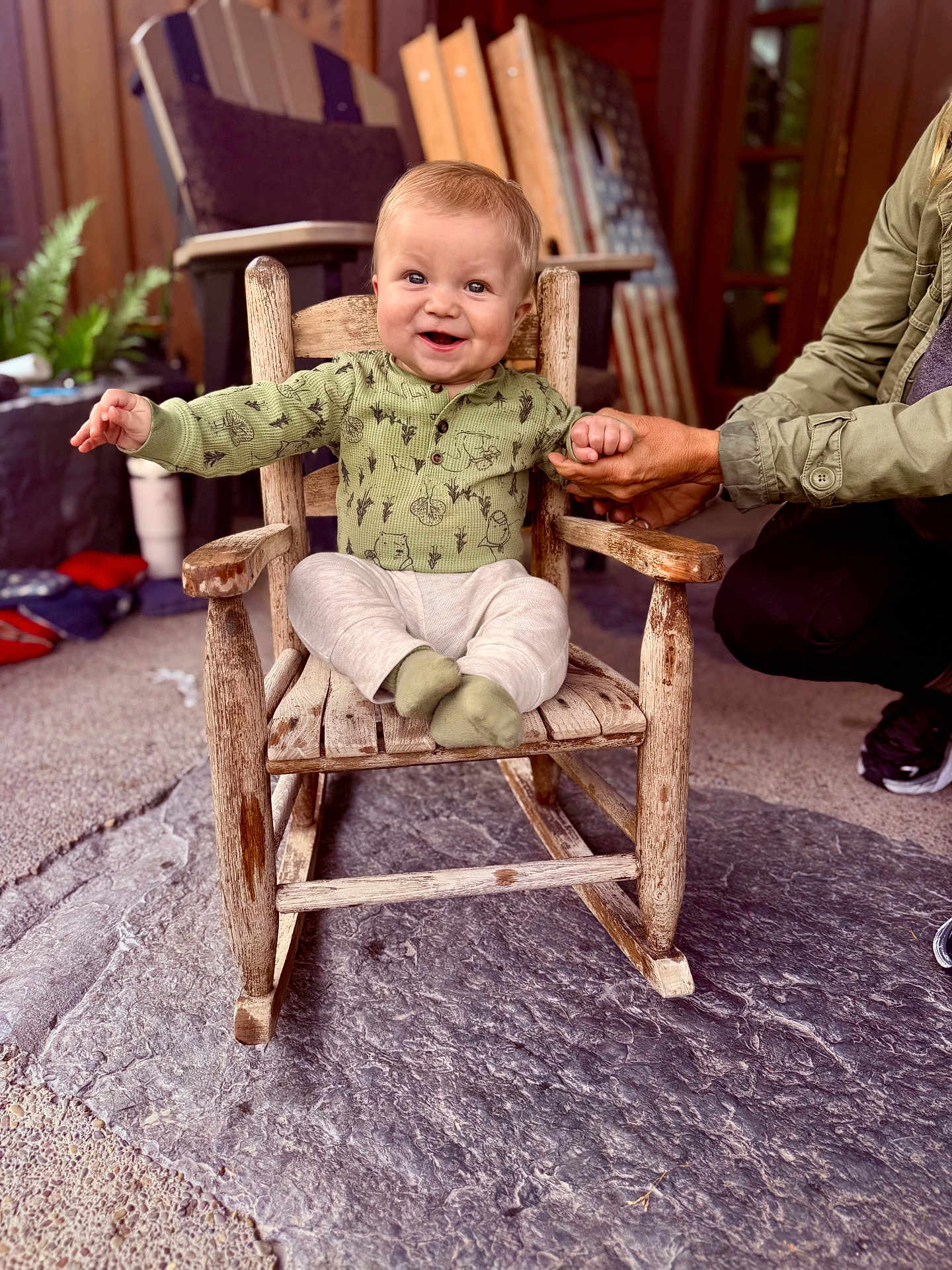 Owen is registered to the contest to win money with this photo: baby, child, rocking_chair, green_shirt, socks, hand_holding, smiling, indoor, wooden_chair, person, adult_hand, floor, plant, flag, carpet, furniture, happy, portrait, cozy, support