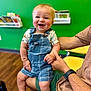 Owen joined the competition — help win amazing prizes! baby, child, smiling, overalls, blue, plaid, person, happy, indoor, green_wall, bookshelf, sitting, adult_hand, holding, shorts, floor, wood_floor, skin, face, portrait