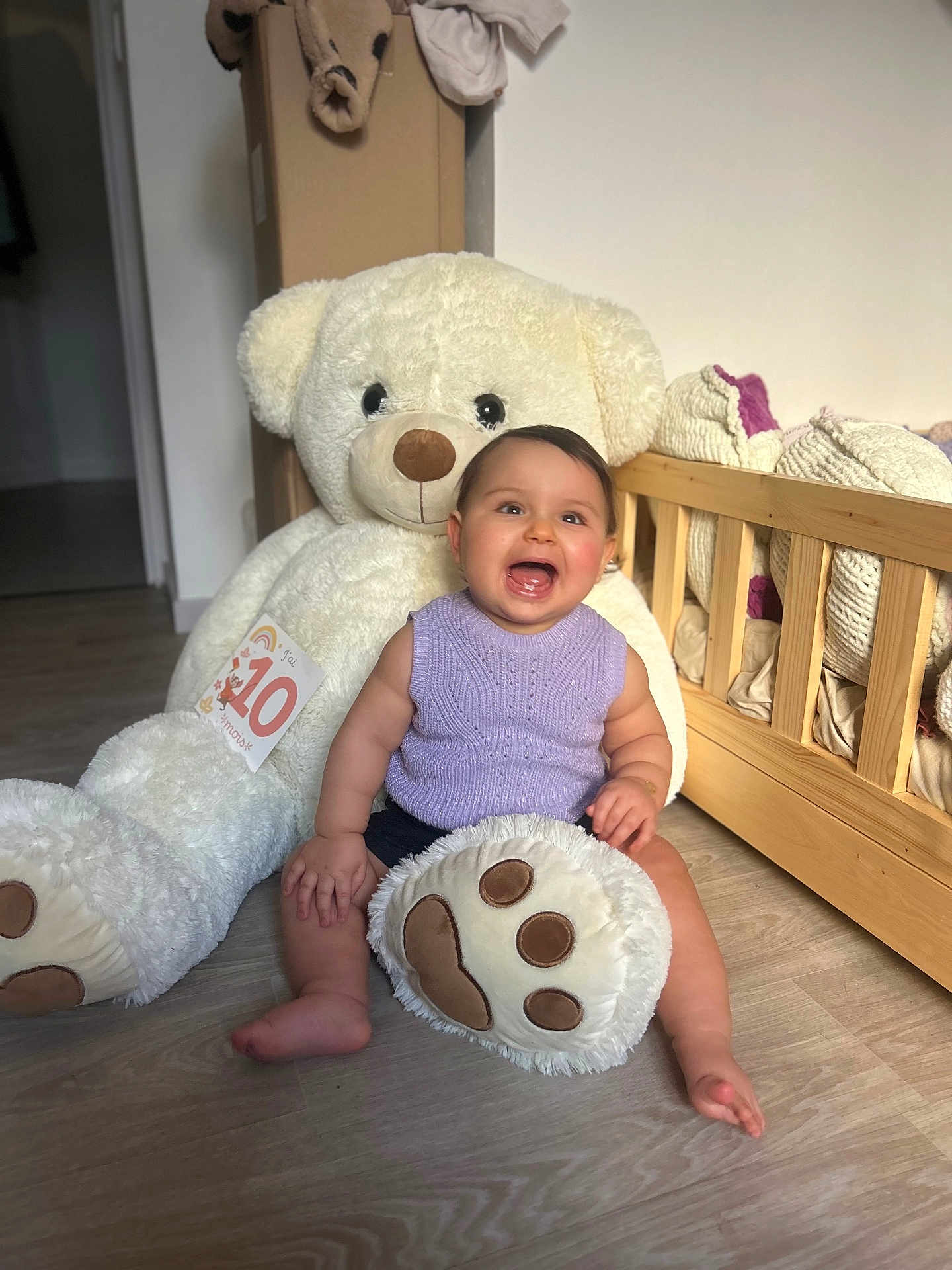 Kayana a rejoint le concours — aidez-le/la à gagner de superbes lots ! baby, smiling_baby, teddy_bear, stuffed_animal, crib, wooden_floor, purple_sweater, sitting, chubby_legs, happy, indoor, plush_paw, milestone_card, cute, portrait, nursery, soft_toy, playful, cozy, child