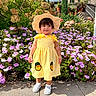child, toddler, portrait, garden, flowers, hydrangea, impatiens, straw_hat, yellow_dress, gingham, polka_dots, outdoor, sunlight, smiling, standing, paved_path, plants, landscaping, name_overlay, fashion