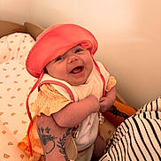 Rose a rejoint le concours — aidez-le/la à gagner de superbes lots ! baby, smiling, hat, coral, knitted_clothing, tattoo, bracelets, arm, holding, nursery, bedding, rainbow_pattern, happy, infant, cute, person, child, indoor, portrait, closeup