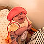baby, smiling, hat, coral, knitted_clothing, tattoo, bracelets, arm, holding, nursery, bedding, rainbow_pattern, happy, infant, cute, person, child, indoor, portrait, closeup