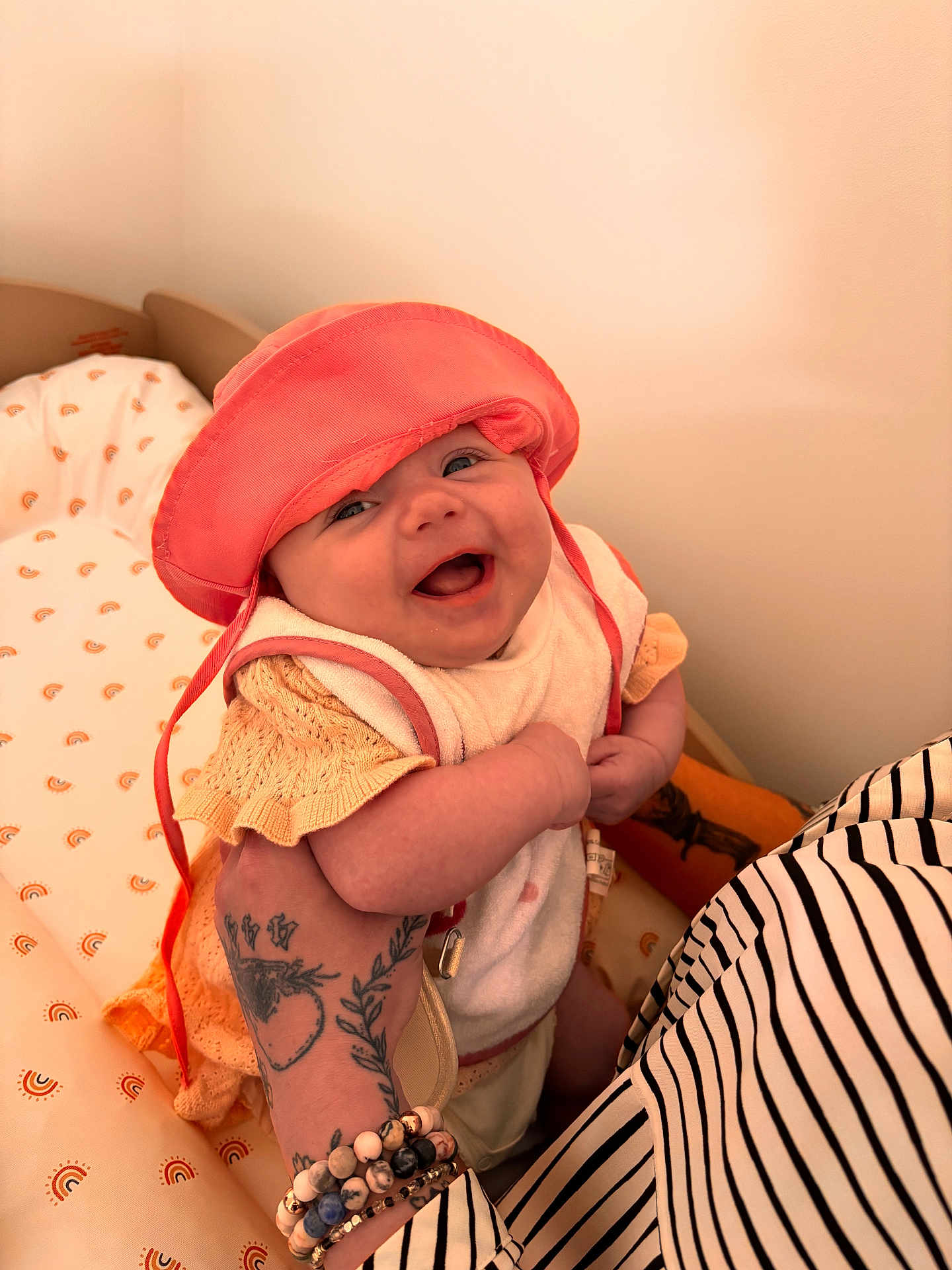 Rose a rejoint le concours — aidez-le/la à gagner de superbes lots ! baby, smiling, hat, coral, knitted_clothing, tattoo, bracelets, arm, holding, nursery, bedding, rainbow_pattern, happy, infant, cute, person, child, indoor, portrait, closeup