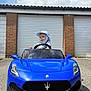 toddler, child, toy_car, blue_car, maserati, steering_wheel, hat, outdoor, garage, brick_wall, pavement, sunny, cloudy_sky, smiling, young_child, vehicle, play, daytime, front_view, casual