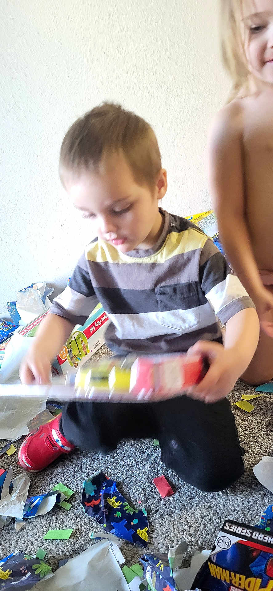 Joseph is registered to the contest to win money with this photo: child, toy, wrapping_paper, mess, carpet, indoor, red_shoes, striped_shirt, play, kids, unwrapping, colorful, floor, person, hands, clothing, room, happy, curly_hair, excitement
