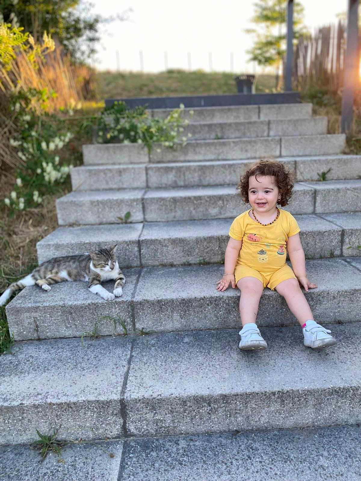 Eva Didar a rejoint le concours — aidez-le/la à gagner de superbes lots ! toddler, child, cat, steps, outdoor, yellow_clothing, smiling, curly_hair, stone, grass, sunlight, happy, sitting, pet, animal, nature, casual, daylight, relaxing, cute
