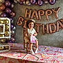 toddler, child, birthday, balloons, chair, table, sticker_book, party, celebration, decorations, number_balloon, floral_tablecloth, rainbow_pattern, wallpaper, smiling, curly_hair, white_dress, socks, indoor, happy