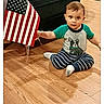 child, toddler, baby, sitting, floor, hardwood_floor, american_flag, chair, striped_pajamas, shirt, socks, indoor, home, furniture, portrait, looking_at_camera, serious_expression, short_hair, photograph, social_media_screen