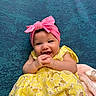 Alaiyah is registered to the contest to win money with this photo: baby, infant, smiling, yellow_dress, pink_headband, clothing, happy, cute, portrait, child, texture, fabric, blanket, pacifier, hand, sitting, indoor, face, person, background