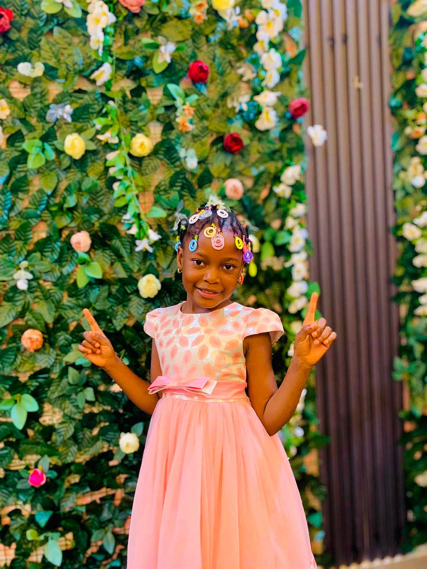Fatim participe au concours pour gagner de l'argent avec cette photo : child, girl, smiling, dress, pink_dress, flowers, flower_wall, greenery, colorful, playful, posing, braids, hair_accessories, outdoor, portrait, happy, fashion, celebration, nature, background