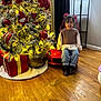 child, smiling, toy_car, christmas_tree, ornaments, presents, holiday, indoors, wooden_floor, curtains, lamp, decorations, festive, girl, ribbons, boots, jeans, sweater, lighting, playful