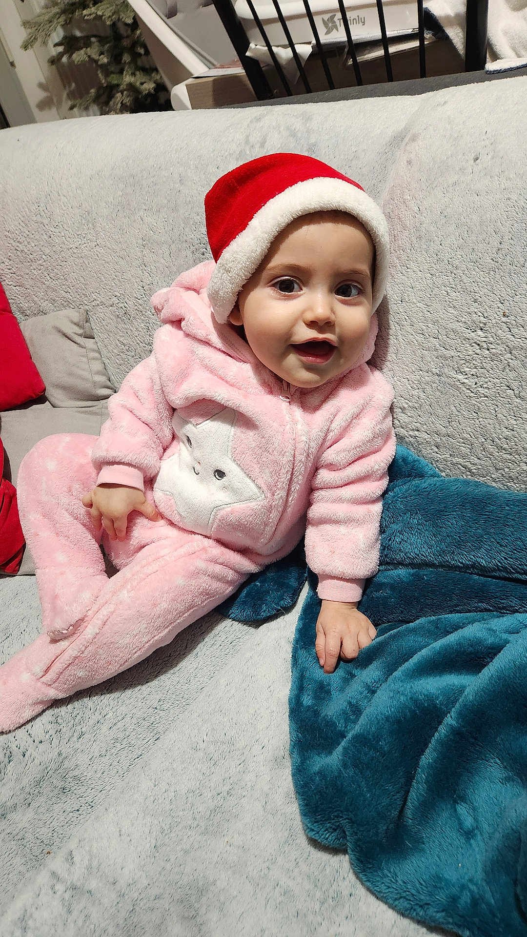 Emma participe au concours pour gagner de l'argent avec cette photo : baby, child, santa_hat, pink_onesie, star_patch, couch, blanket, red_cushion, gray_couch, smiling, indoor, holiday, festive, plush, soft_texture, happy, portrait, seated, young_child, cozy