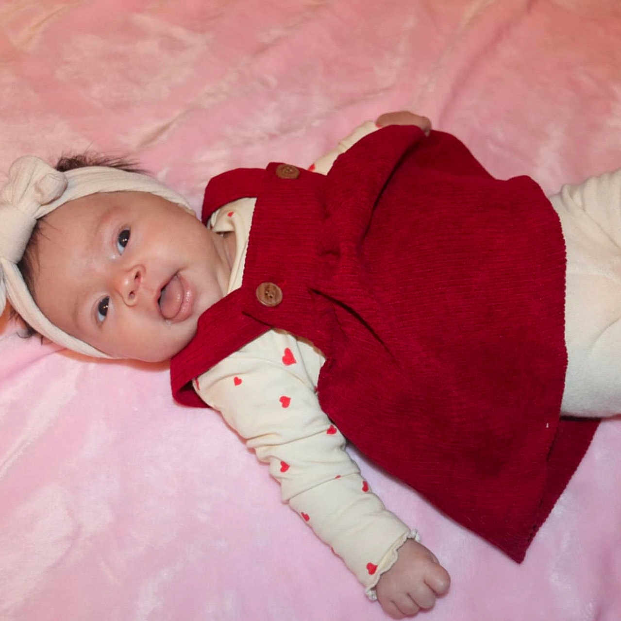 Romy a rejoint le concours — aidez-le/la à gagner de superbes lots ! baby, button, child, cozy, cute, face, happy, headband, heart_pattern, infant, lying_down, newborn, onesie, pink_blanket, portrait, red_dress, smile, socks, soft_texture, tongue_out