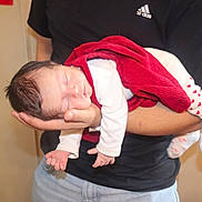 Romy a rejoint le concours — aidez-le/la à gagner de superbes lots ! baby, newborn, sleeping, red_blanket, white_clothing, heart_pattern, socks, adult_arm, holding, indoor, person, tender_moment, peaceful, infant, skin, head, hand, black_shirt, jeans, human