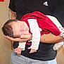 baby, newborn, sleeping, red_blanket, white_clothing, heart_pattern, socks, adult_arm, holding, indoor, person, tender_moment, peaceful, infant, skin, head, hand, black_shirt, jeans, human