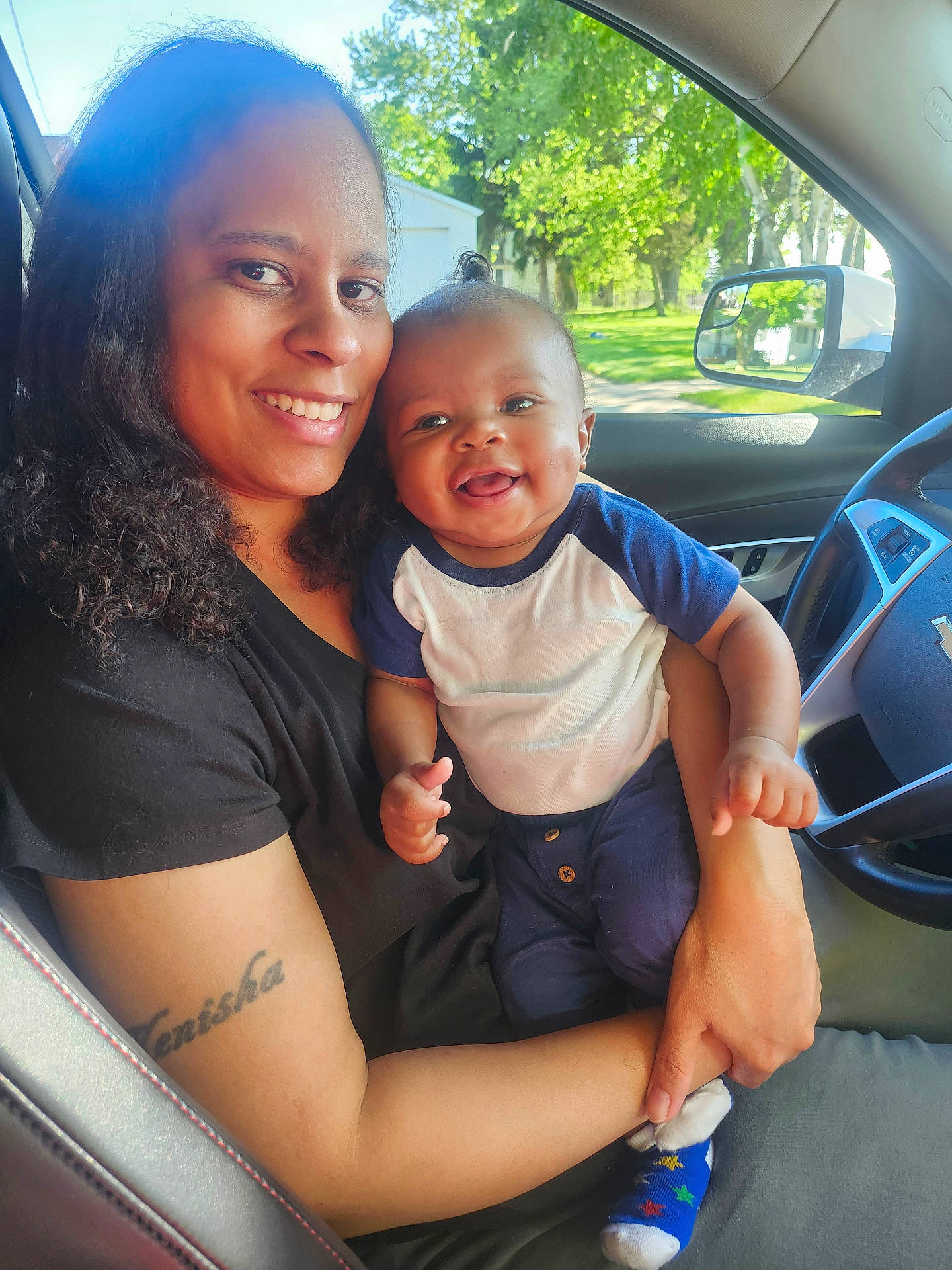 Alexander is registered to the contest to win money with this photo: baby, car_interior, casual, clothing, daylight, greenery, happy, holding, infant, nature, person, portrait, seat, smile, socks, steering_wheel, sunlight, tattoo, window, woman