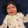 baby, child, infant, smiling, happy, portrait, close_up, face, bonnet, headwear, pajamas, cherry_pattern, clothing, lying_down, indoor, dark_background, blue_blanket, toothless_smile, baby_fashion, adorable