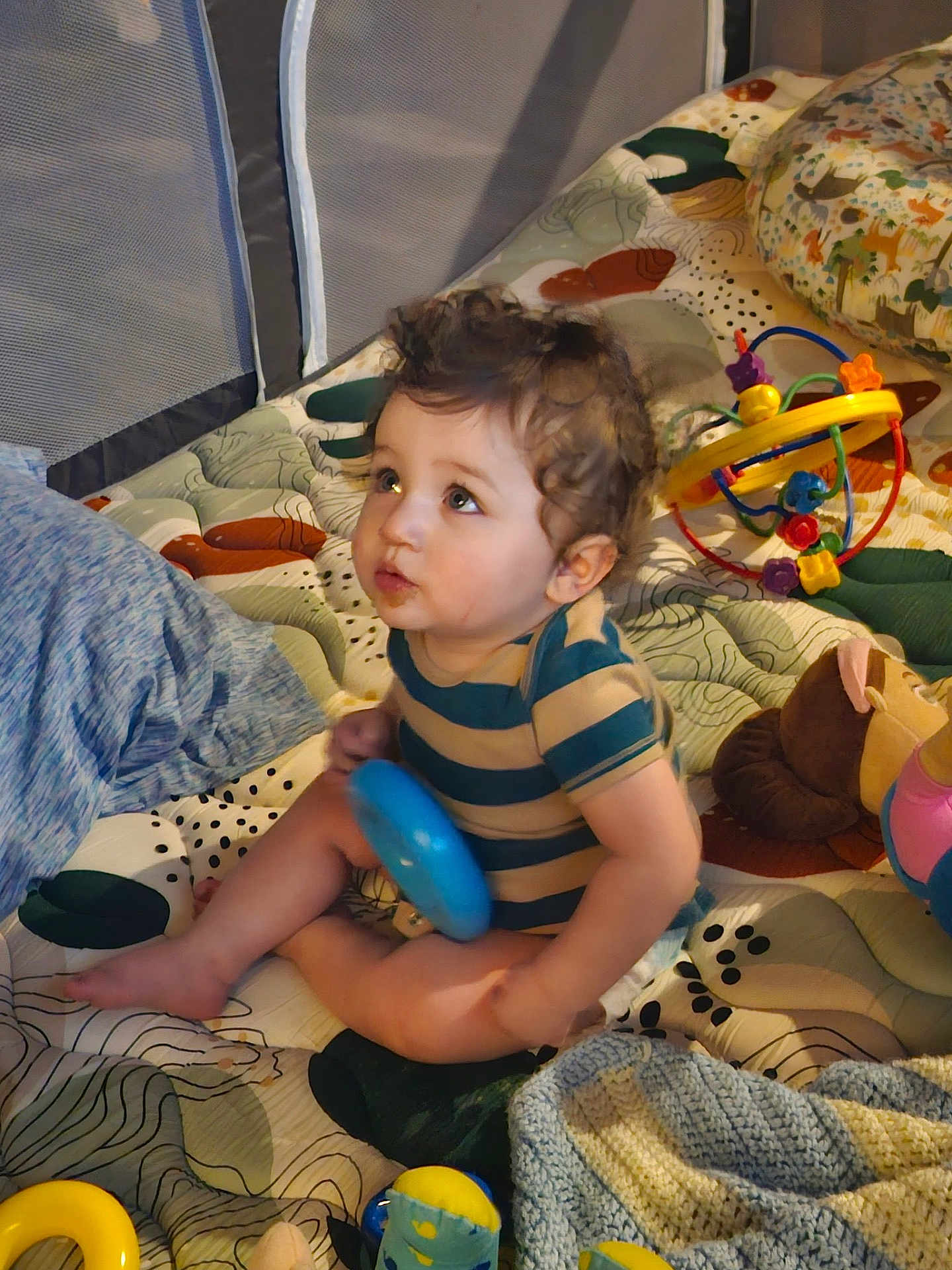 Kai is registered to the contest to win money with this photo: baby, child, curly_hair, striped_shirt, toy, blanket, playpen, indoor, sitting, looking_up, colorful, patterned, soft_toys, infant, cute, young_child, floor_play, multicolored, happy, small_person