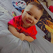 Mateo is registered to the contest to win money with this photo: baby, smiling, red_shirt, bed, pillow, blanket, indoor, child, happy, face, person, cute, plush, comfort, lying_down, young_child, cozy, soft, head, infant