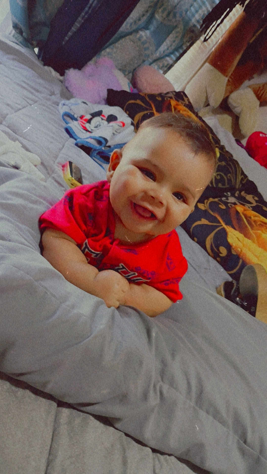 Mateo is registered to the contest to win money with this photo: baby, smiling, red_shirt, bed, pillow, blanket, indoor, child, happy, face, person, cute, plush, comfort, lying_down, young_child, cozy, soft, head, infant