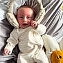 baby, infant, earmuffs, plush_toy, daisy, bed, blanket, sleepwear, soft_texture, cute, child, smile, face, hand, lying_down, cozy, indoor, comfort, toy, expression