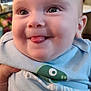 baby, child, tongue_out, smiling, blue_shirt, dinosaur_print, face, eyes, cheeks, indoor, person, cute, infant, expression, happy, closeup, skin, hand, sitting, young