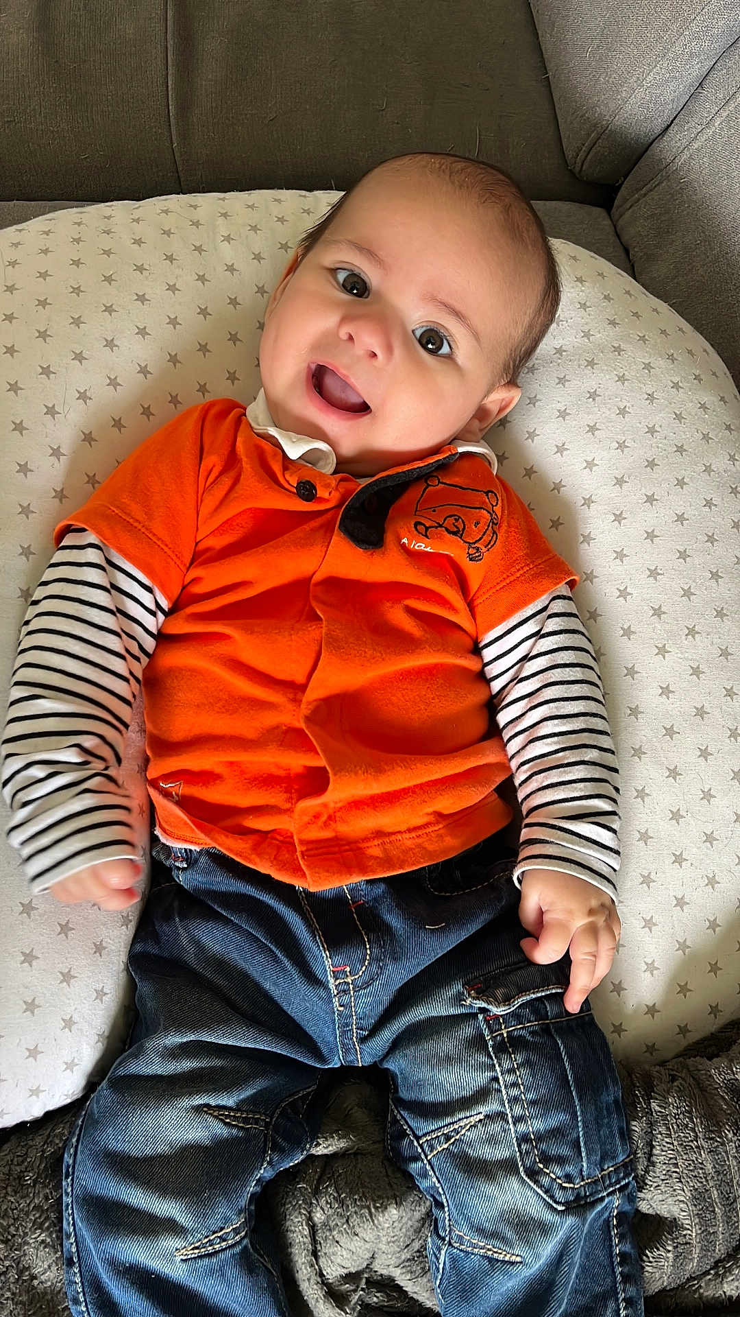Hakan a rejoint le concours — aidez-le/la à gagner de superbes lots ! baby, child, smiling, orange_shirt, striped_sleeves, blue_jeans, cushion, star_pattern, face, happy, indoors, seated, clothing, person, cute, young_child, portrait, jeans, long_sleeve, comfort