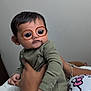 adult_hand, baby, bracelet, cartoon_eyes, child, cute, expression, face_filter, glasses, green_clothing, hand, hello_kitty, holding, indoor, person, playful, portrait, shirt, skin, wall