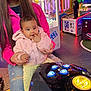 Sonia joined the competition — help win amazing prizes! baby, child, woman, arcade, game_machine, colorful_lights, pink_jacket, ripped_jeans, thumb_in_mouth, indoor, playful, smile, young_boy, standing, clothing, entertainment, fun, people, casual, family