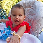 Sonia is registered to the contest to win money with this photo: baby, child, high_chair, toy, outdoor, greenery, bicycle, red_clothing, smiling, happy, infant, playful, cushion, person, portrait, sunlight, nature, cute, young, reaching