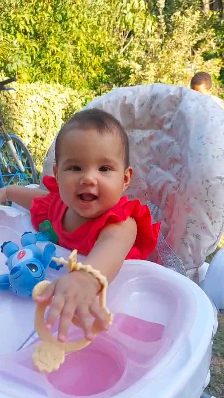Sonia is registered to the contest to win money with this photo: baby, child, high_chair, toy, outdoor, greenery, bicycle, red_clothing, smiling, happy, infant, playful, cushion, person, portrait, sunlight, nature, cute, young, reaching