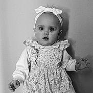Miley is registered to the contest to win money with this photo: baby, child, girl, headband, bow, dress, floral_pattern, long_sleeves, portrait, monochrome, black_and_white, standing, curious_expression, indoors, wall, hands, face, cute, young, person