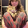 person, cat, plaid_pants, long_hair, indoor, carpet, bracelet, fluffy_cat, winking_cat, sitting, holding, smiling, black_shirt, home, door, furniture, relaxed, cute, cozy, pet