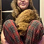 person, cat, indoor, plaid_pants, brown_cat, hair, smile, sitting, hugging_knees, floor, bracelet, furniture, carpet, cozy, pet, animal, casual_clothing, relaxed, comfort, home