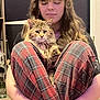 person, cat, plaid_pants, long_hair, indoor, sitting, fluffy_cat, crossed_legs, bracelet, carpet, guitar, glassware, casual, relaxed, brown_hair, tabby_cat, holding, home, pet, cozy