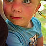 child, toddler, portrait, close_up, face, blue_eyes, blond_hair, serious_expression, side_eye, outdoor, sunlight, colorful_shirt, blue_clothing, sitting, person, family, candid, photography, background_blur, daylight