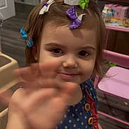 Evie is registered to the contest to win money with this photo: toddler, child, girl, hair_clips, smile, hand, indoor, toy, furniture, dress, polka_dot, wooden_floor, pink_chair, buttons, playroom, cute, happy, person, face, waving