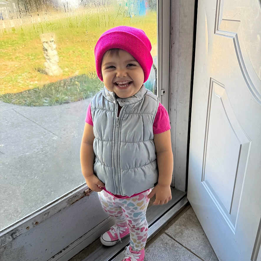 Evie joined the competition — help win amazing prizes! beanie, casual_clothing, child, cute, daylight, door, floor_tile, glass, happy, heart_pattern, indoor, leggings, person, pink, smiling, sneakers, standing, toddler, vest, window