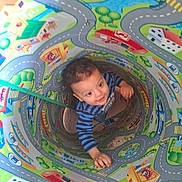 Jasper is registered to the contest to win money with this photo: toddler, child, play_tunnel, colorful, road_pattern, buildings, trees, curious, indoor, toy, playtime, striped_shirt, young_child, smiling, vibrant_colors, cartoon, cityscape, hand, looking_up, cute