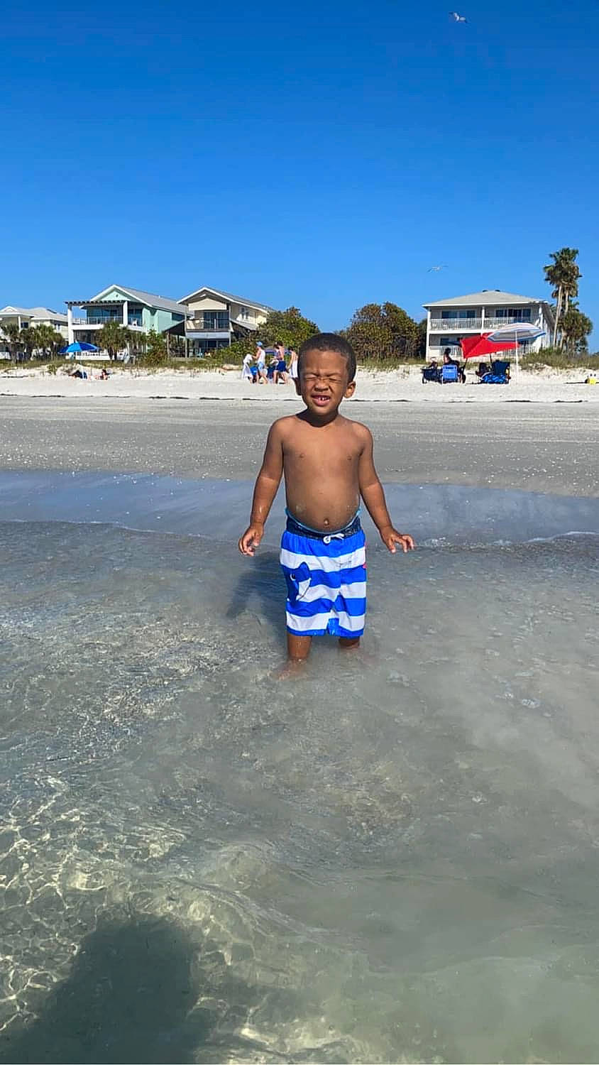 Christopher is registered to the contest to win money with this photo: azure, barechested, beach, board_short, body_of_water, daytime, electric_blue, fun, happy, leisure, people_on_beach, person, sand, shore, sky, toddler, travel, tree, trunk, trunks