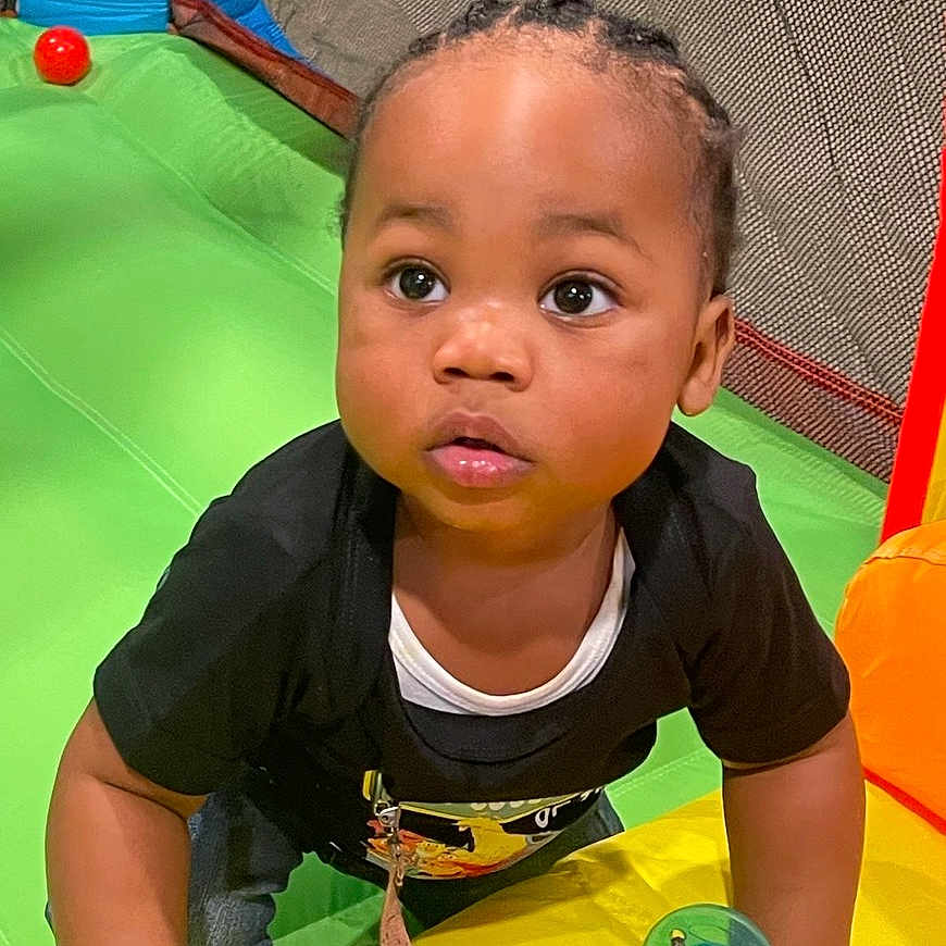 Khyren is registered to the contest to win money with this photo: toddler, child, baby, portrait, indoors, playground, play_structure, colorful, climbing, sitting, looking_at_camera, wide_eyes, curly_hair, braids, black_shirt, pacifier, green_slide, yellow_surface, red_ball, play_area