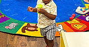 Khyren joined the competition — help win amazing prizes! child, toddler, standing, playing, toy, playroom, colorful, alphabet_rug, braids, shorts, sandals, indoors, daycare, furniture, activity_table, plastic_toy, green_toy, curious, portrait, wooden_floor