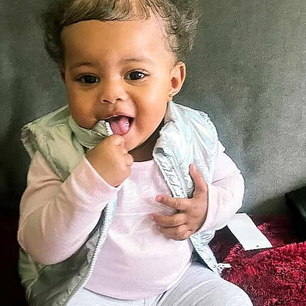 Carebear is registered to the contest to win money with this photo: toddler, child, smiling, sitting, indoors, clothing, vest, long_sleeve, pants, hair_tie, curly_hair, cute, playful, face, hand, blanket, fabric, portrait, happy, young_child
