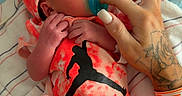 Oakley is registered to the contest to win money with this photo: newborn, baby, infant, hospital, hospital_bed, pacifier, red_outfit, hat, cap, blanket, bedding, hand, tattoo, adult, caregiver, portrait, lying_down, indoor, medical_wristband, family